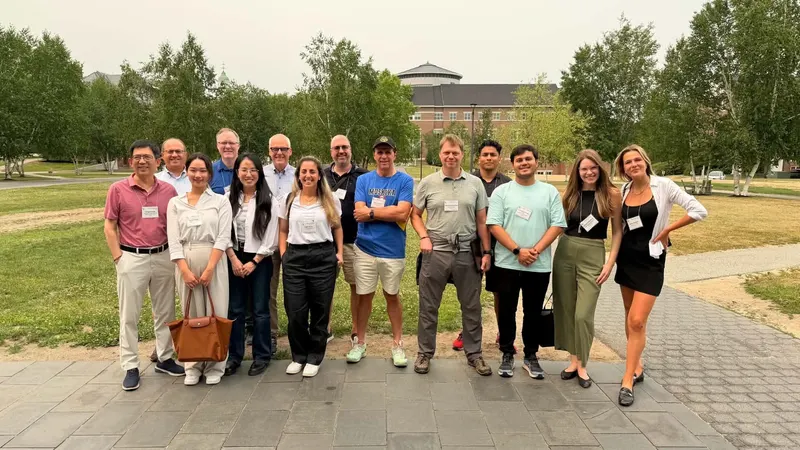 A large group of conference attendees posing together outdoors on a paved pathway with campus buildings and trees in the background.