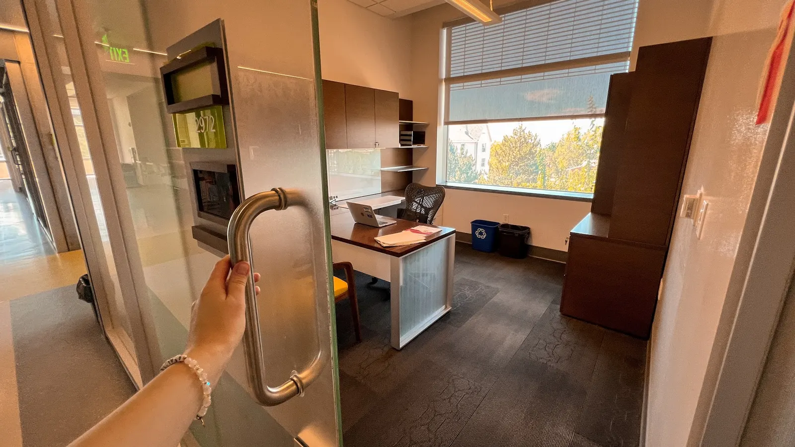 A person's hand opens a glass door revealing a modern, sunlit office with a wooden desk, office chair, computer, and a large window looking out onto trees.