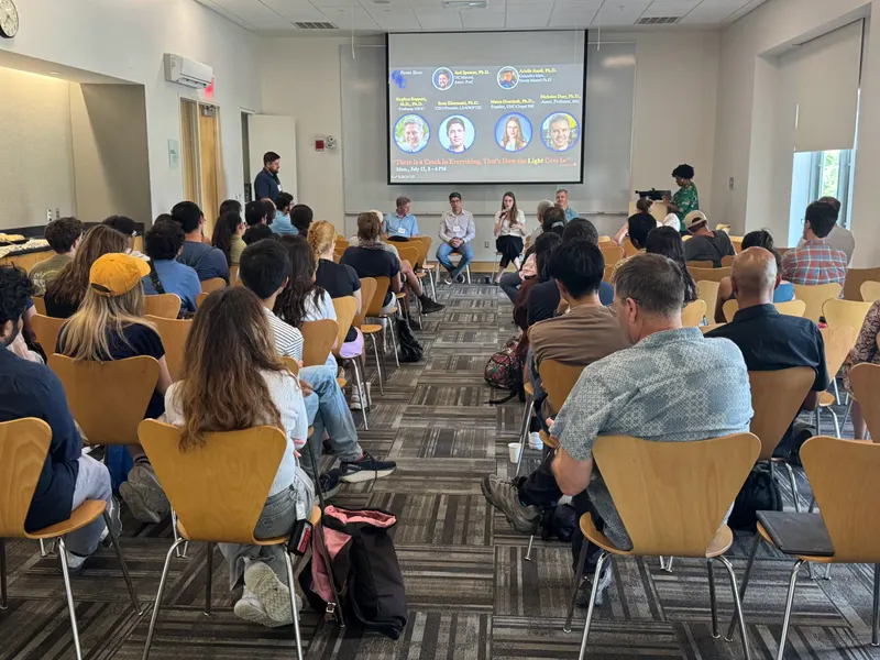 A panel presentation taking place at Gordon Research Conference with a large audience seated in chairs facing a projection screen displaying speaker profiles.