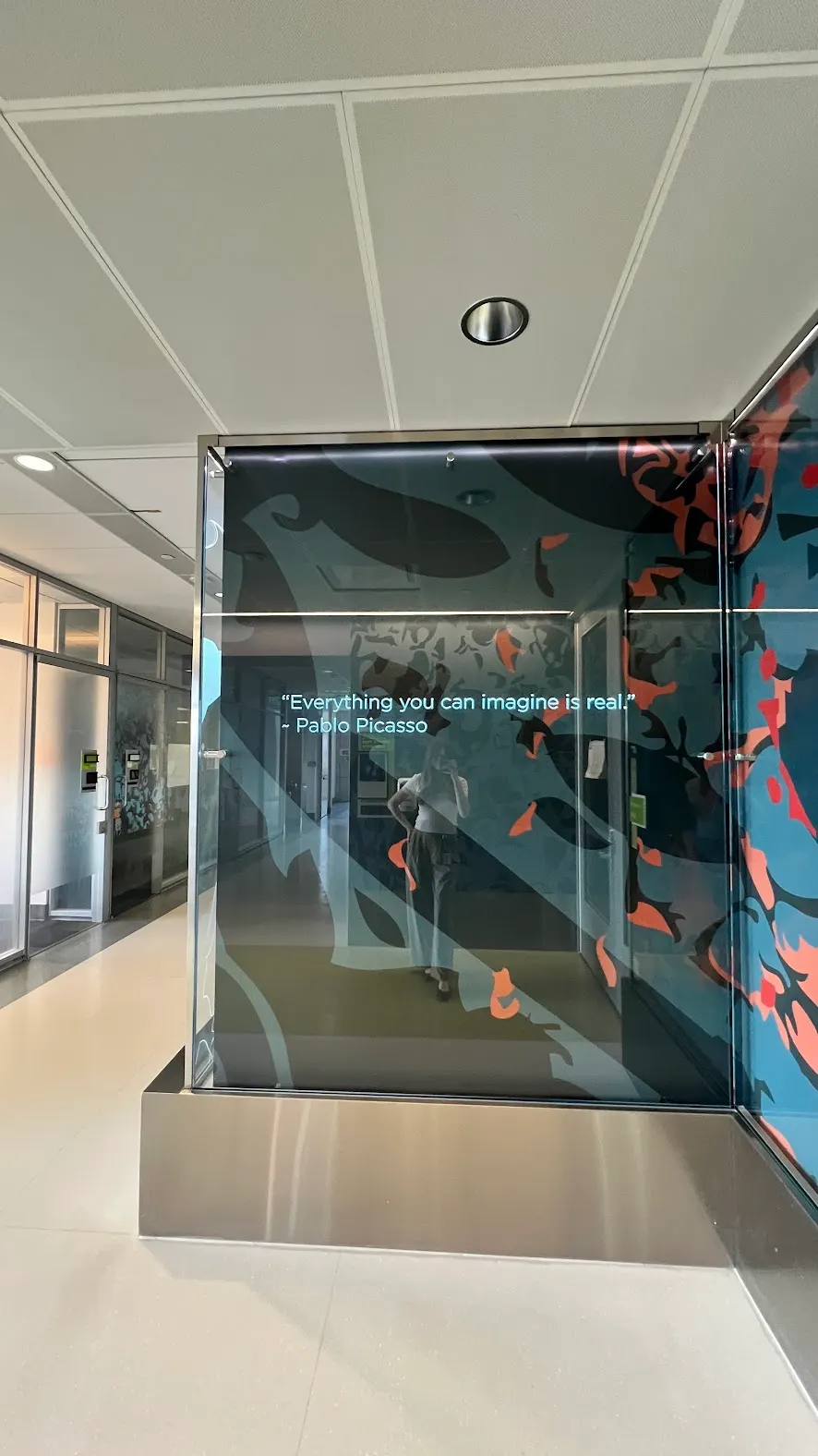 A glass feature wall in a modern building hallway displaying the Pablo Picasso quote, "Everything you can imagine is real," with a reflection of a person taking the photo.