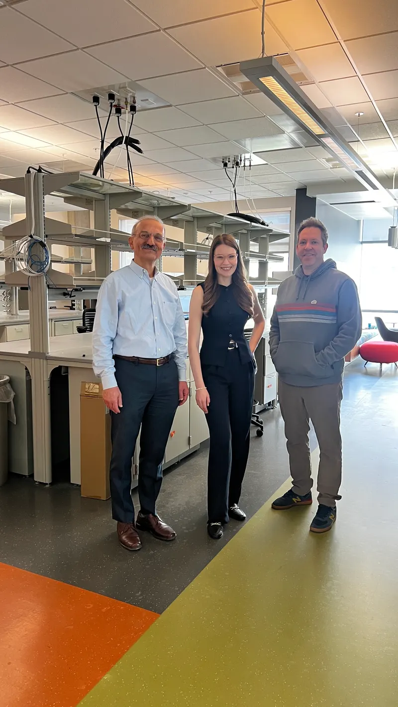 Marta, Shawn, and Hamid posing together and smiling inside a research laboratory with equipment benches in the background.