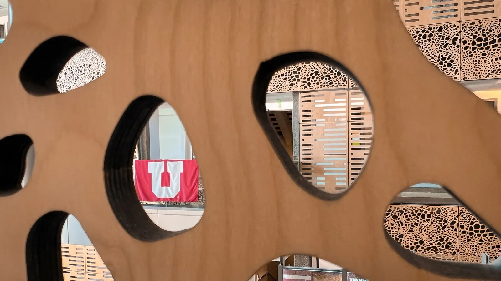 A view through an artistic wood cutout panel featuring rounded openings, revealing a University of Utah flag and textured interior architecture in the background.