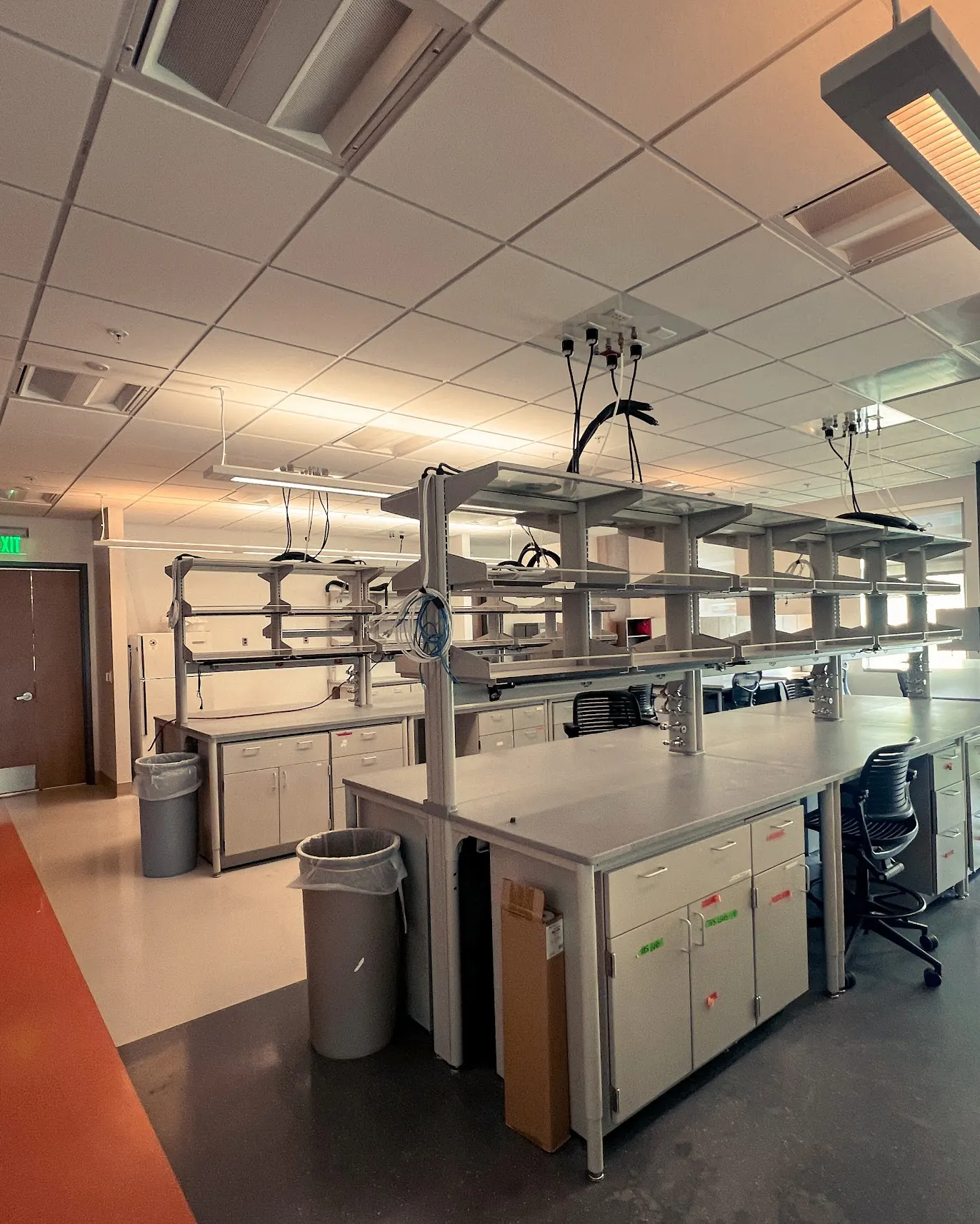 A bright, spacious, and empty research laboratory with modern workbenches, overhead shelving, gas/utility connections, storage cabinets, and rolling lab chairs.