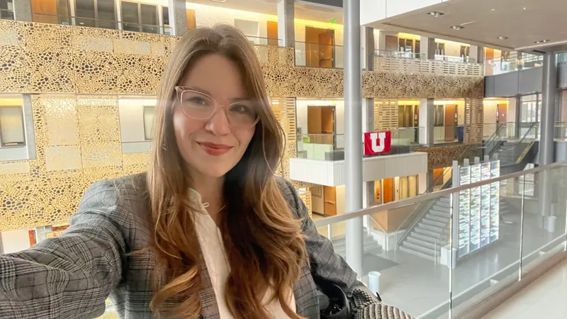 A woman taking a selfie inside the Skaggs research building. A modern multi-story atrium with glass railings, patterned wall panels, and a University of Utah flag in the background.